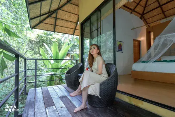 Woman relaxing on a lodge balcony beside a bed with mosquito netting, looking over rainforest greenery at Khao Sok National Park.