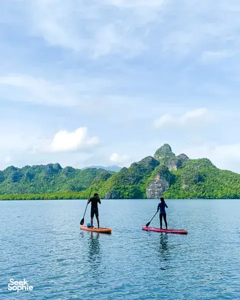 两位立式桨板体验者站在平静的水面上,身后是葱郁的山丘和悬崖,拍摄于丹绒鲁的 SUP 探险途中
