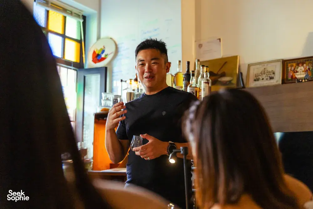 Host explaining flavours during a small-batch whisky tasting in Singapore, reaching up a bottle as guests listen in a home bar