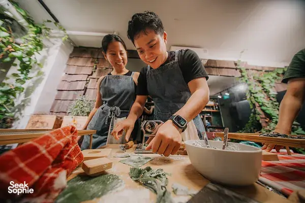 Guests arranging herbs and rolling pasta dough on a wooden table in a leafy kitchen in Singapore during workshops in Singapore