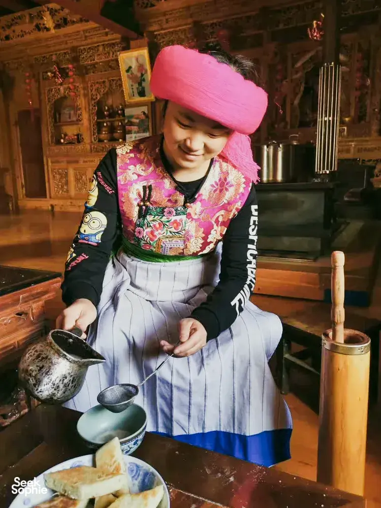 Woman in a pink headscarf pouring tea from a small kettle at a traditional Tibetan home visit in Shangri-La