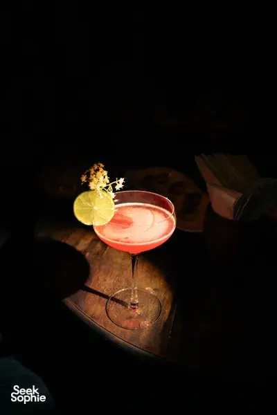 傍晚昏暗中，会安一张木桌上的一杯红色鸡尾酒，配有青柠片和可食用花朵装饰