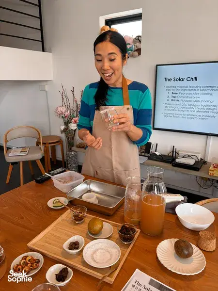 The TCM Chef smiling while holding a small glass of layered mocktail, with a learning screen behind, in workshops in Singapore
