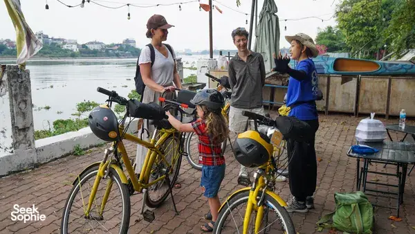 Travellers on yellow bikes pause by the riverside on a Mekong Island trip from Ho Chi Minh City, with a boat behind