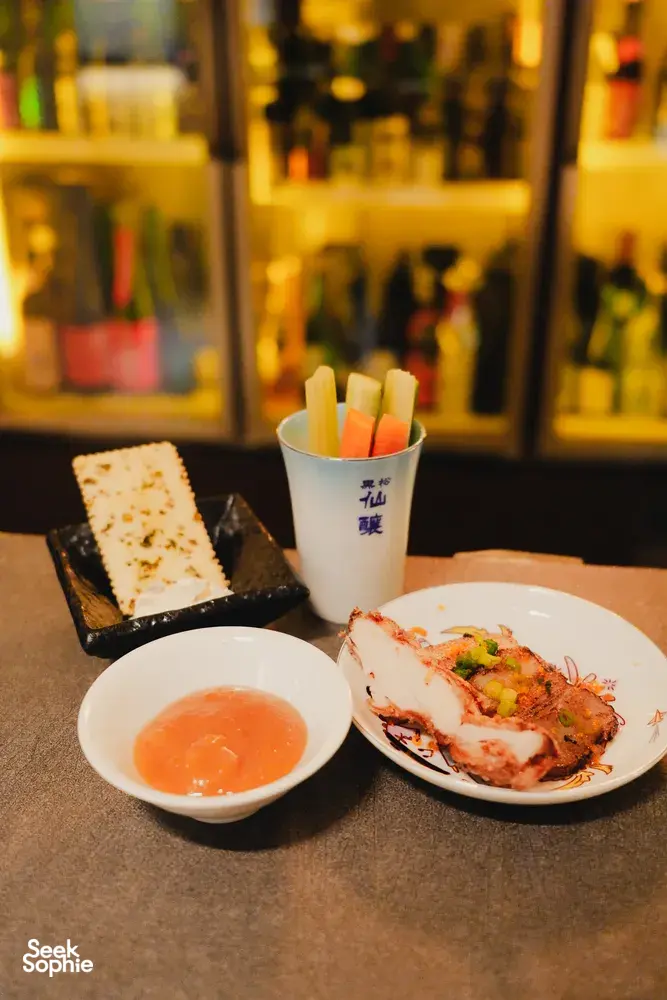 Savoury appetisers and sake tasting cups on a table in a tucked-away Singapore shop, with bottles lining the bar