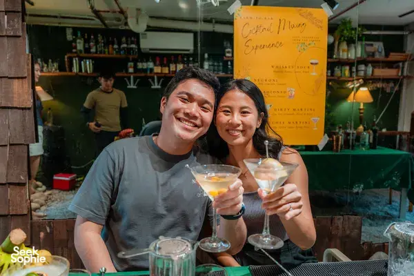 Two people smile and clink cocktail glasses on a bar table during a private cocktail making class in Singapore’s Cosy Nature Hideout
