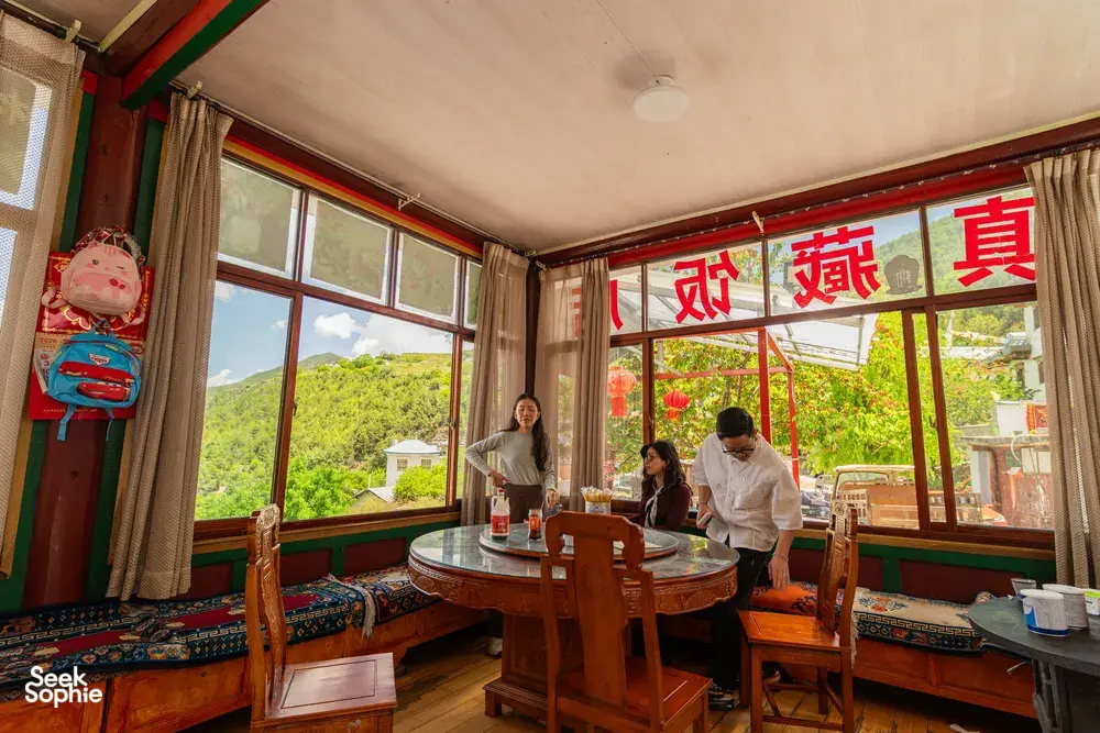 Lodge dining room in Shangri-La with large windows to green hills, travellers at a round table preparing snacks