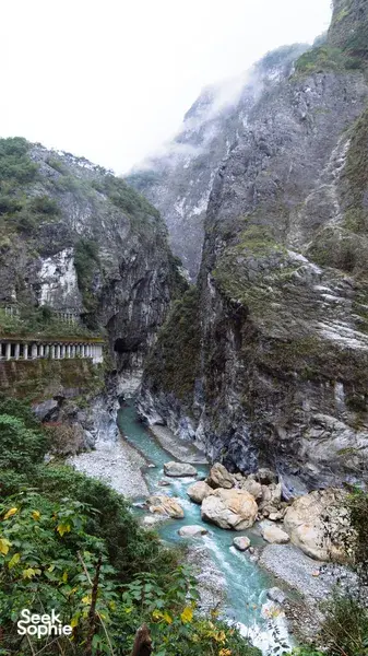 Narrow mountain gorge trail with a clear blue stream cutting between steep rock cliffs and a riverside bridge in China