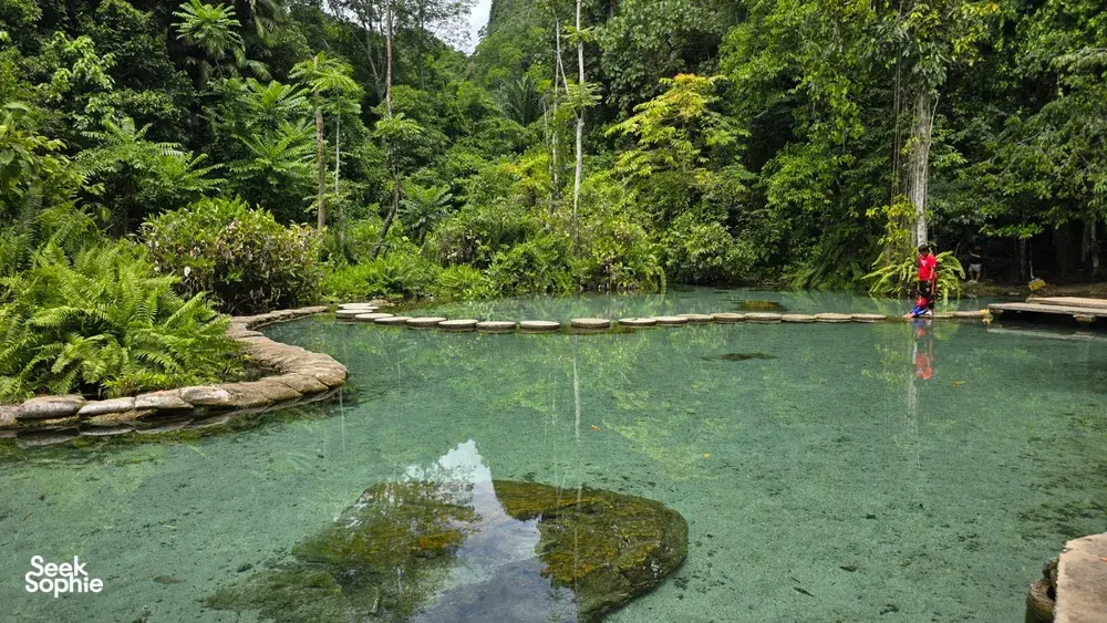 People relax and wade in shallow turquoise Blue Spring water next to floating rafts, surrounded by rainforest in Khao Sok National Park