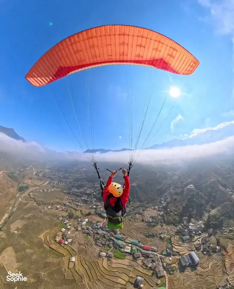 Paragliding rider in a red jacket reaching up beneath an orange canopy over terraced valley slopes, Sapa