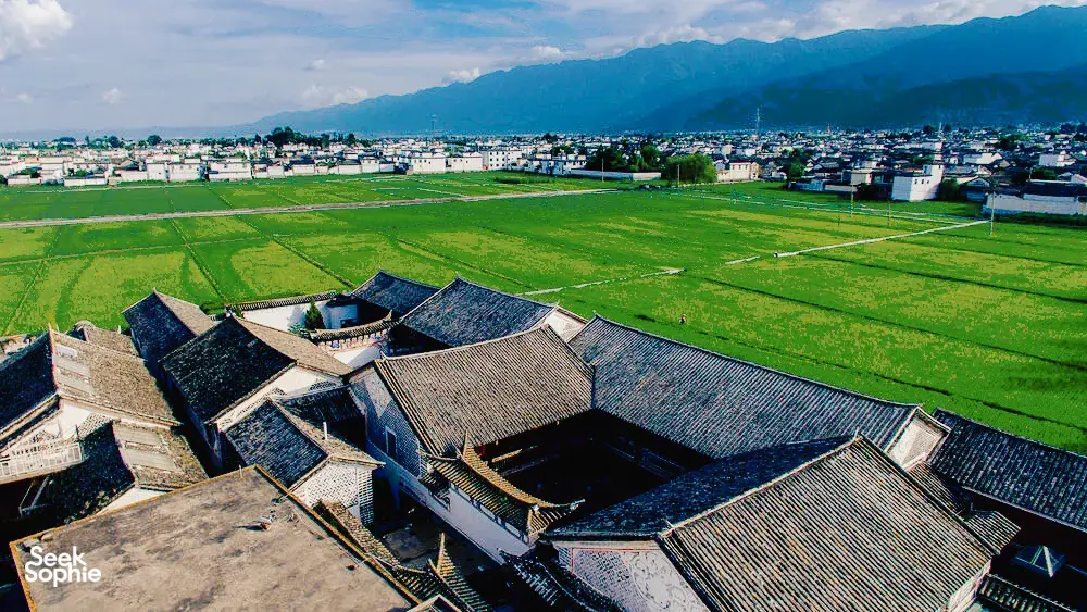 Pemandangan udara rumah-rumah beratap genting di pekan lama Xizhou, Dali, dikelilingi kehijauan sawah dan banjaran gunung di kejauhan