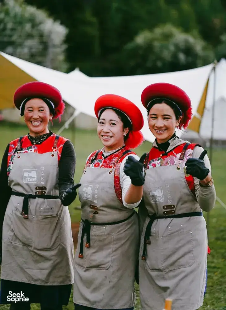 Three women in traditional Tibetan-style outfits and red fur hats smiling at a camp in Shangri-La’s remote valleys
