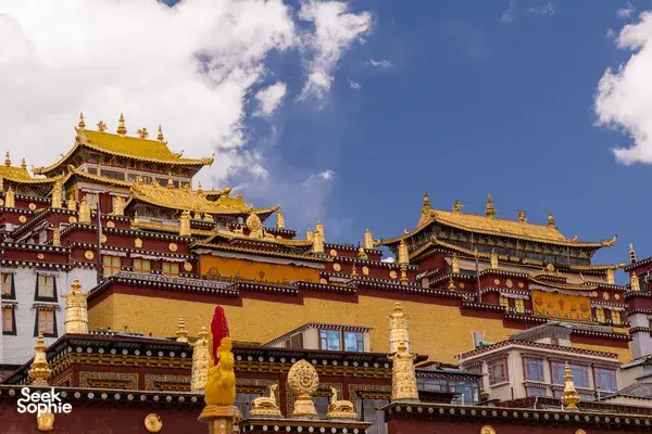 Ornate golden-roofed Tibetan-style palace buildings in Shangri-La against a bright blue sky, with scattered clouds overhead