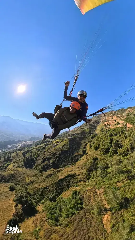 Tandem paragliding above grassy mountain ridges, with two orange-helmeted riders soaring against blue sky in Sapa