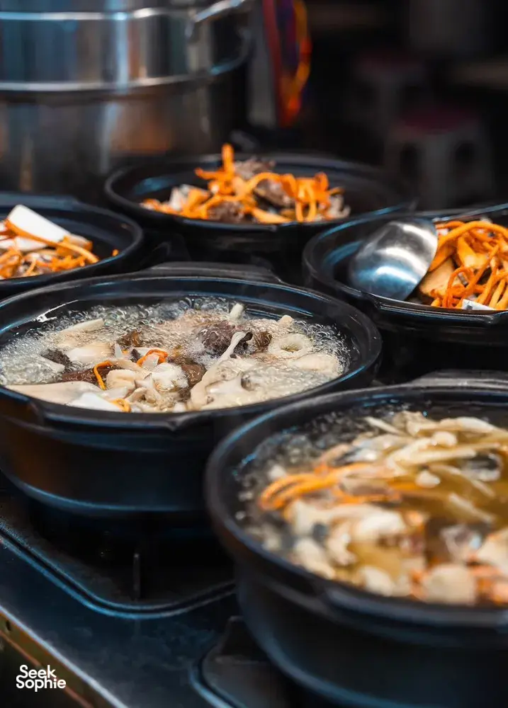 Hotpots simmering in metal pans with vegetables and fish for tasting at a local Lijiang street food stop