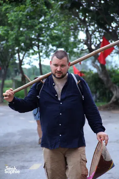Doai Tour & Homestay host carrying a basket and hoe outdoors along a rural path during hands-on rice farming near Hanoi