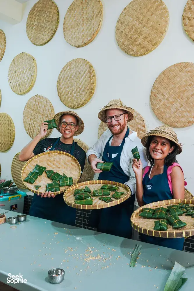 Small group in aprons and hats during a Bali tempe making workshop, holding woven trays with leaf-wrapped tempe