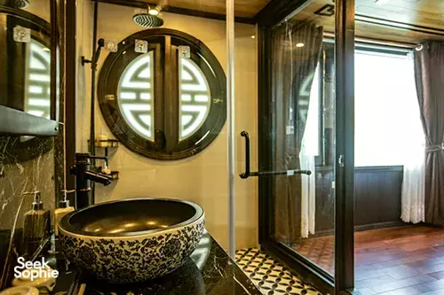Cat Ba Ocean Cruises ensuite bathroom with a decorative washbasin, round mirror and shower area on the wooden boat in Halong Bay