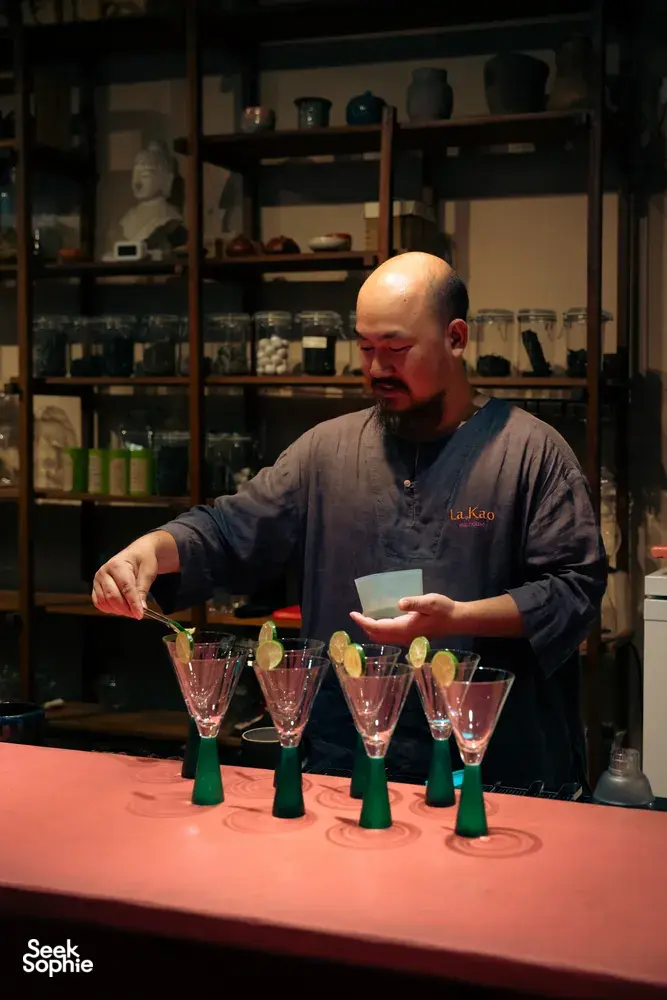 Bartender menuang minuman dari botol hijau untuk para tetamu, dengan gelas koktel dan hiasan di sebuah hentian merasa di Hoi An