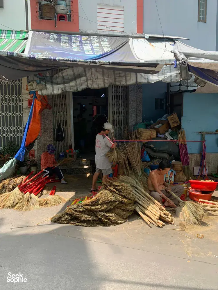 Straw brooms and bundles on the ground outside a Saigon craft alley workshop, where an artisan tends materials by the door