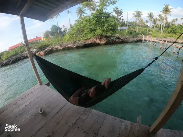 Man relaxing in a hammock on the wooden deck of a waterfront sea lodge, looking out over calm green water in Semporna