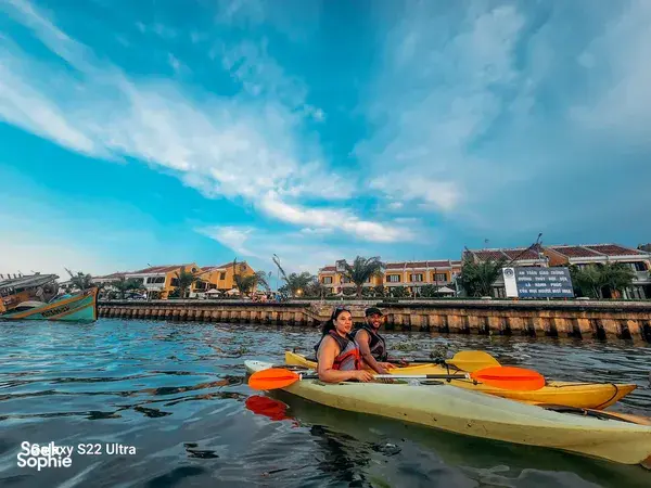 Two kayakers in bright paddles sit near the Hoi An waterfront promenade, sheltered by buildings as they float on calm water