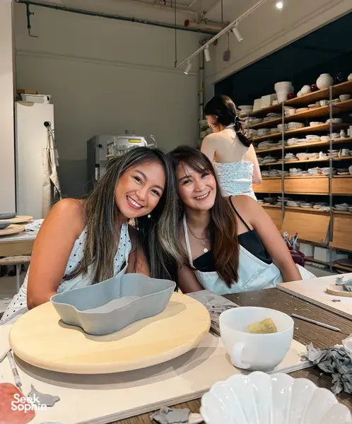 Two friends smiling at a Singapore pottery studio table with European-style ceramic plates and bowls drying behind them