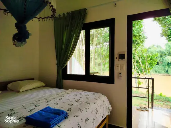 Green Hope Lodge bedroom corner with patterned duvet and green curtains, looking out from the terrace to rainforest greenery at Cat Tien