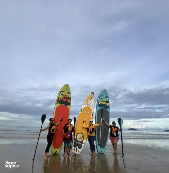 Paddleboarders stand in the shallows with bright boards and paddles on a wide sandy beach near Kota Kinabalu