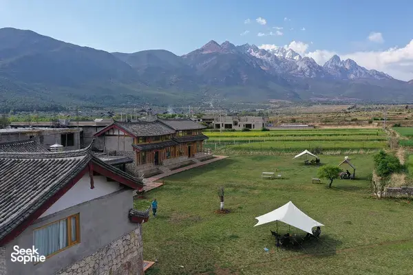 Lijiang coffee estate courtyard: traditional timber buildings, white tents on a green lawn, and misty mountains beyond
