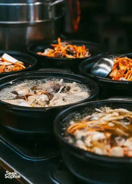 Rows of black soup pots at a Lijiang food stall with bubbling broth, fish pieces, and orange shredded vegetable topping