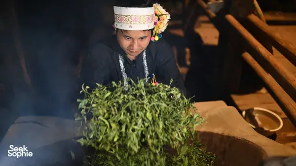 Pembuat teh tradisional Xishuangbanna sedang bekerja dengan tangan di meja kayu, memakai ikat kepala bercorak sambil memproses daun teh