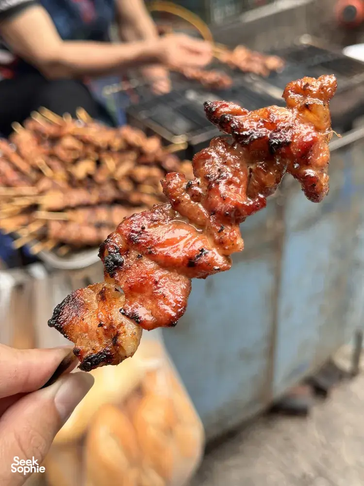 Hand holding char-grilled pork pieces over a hot barbecue grill during a Hanoi food adventure