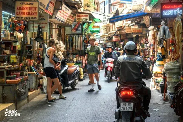 Cyclists riding a scooter through a busy shop-lined alley in Hanoi, with street stalls and pedestrians on both sides