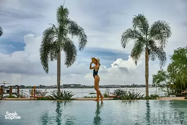 Guest standing in a rooftop-style pool facing a wide Mekong Delta river, framed by palm trees at The Island Lodge