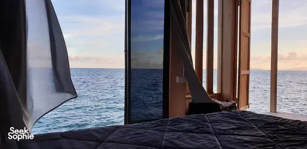 Sea lodge cabin bed by the window with curtains open, facing calm ocean off Semporna during evening