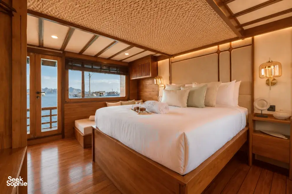 Sunlit phinisi cabin interior with a wooden-framed double bed, layered bedding and bedside lamps, overlooking the ocean in Labuan Bajo