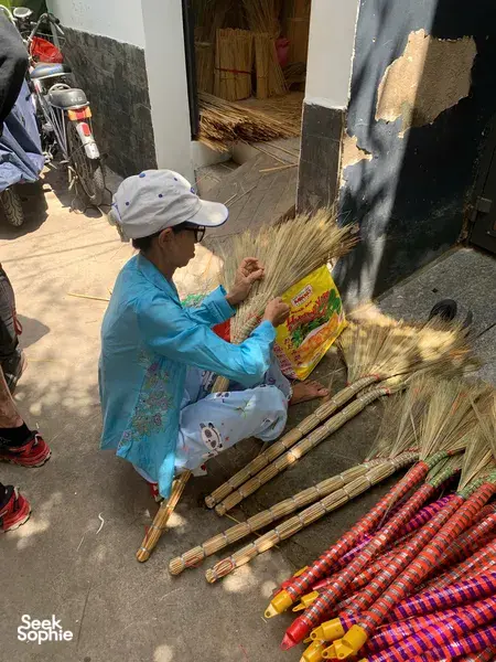 Saigon artisan in a straw hat weaving and packing broom brushes, with rows of finished thatch brooms laid out nearby