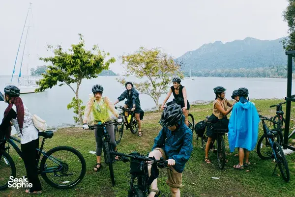 Bicycle Day Langkawi cyclists pause by the bay on Langkawi, standing with bicycles near calm water and distant hills