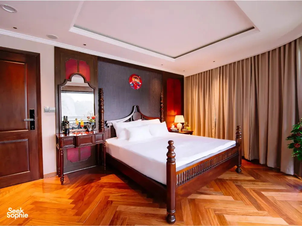 Elegant Cat Ba cruise suite bedroom with large bed, decorative wall mirror, vanity with sink and warm golden lighting