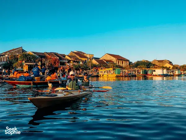 Paddlers in kayaks on the Hoi An river, with warm-lit riverside houses and a wooden walkway along the harbour at sunset