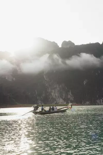 Longtail boat cruising on misty lake in Khao Sok with orange life jackets, backed by cliffs — Khao Sok National Park