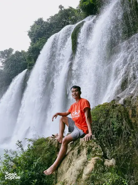 Solo traveller sitting on a grassy cliff beside a waterfall in Cao Bang on a Ha Giang Expeditions motorbike loop
