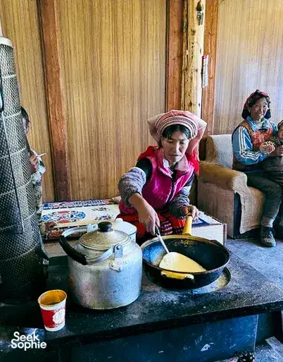 Lijiang village kitchen where a local woman cooks by a stove, with another person seated nearby on a day meal