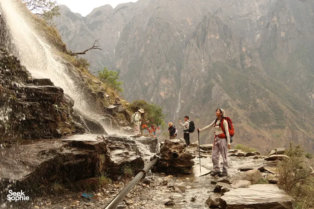 Hikers on the Tiger Leaping Gorge high trail in Lijiang, wading along a rocky route beside a roaring waterfall