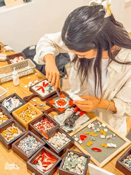 Participant placing ceramic mosaic tiles inside a wooden tray at Bat Trang, Hanoi, leaning over the work in progress