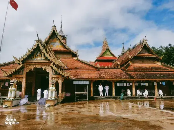 Ornate temple buildings in Xishuangbanna with gilded roofs, statues and a rain-wet courtyard under cloudy skies