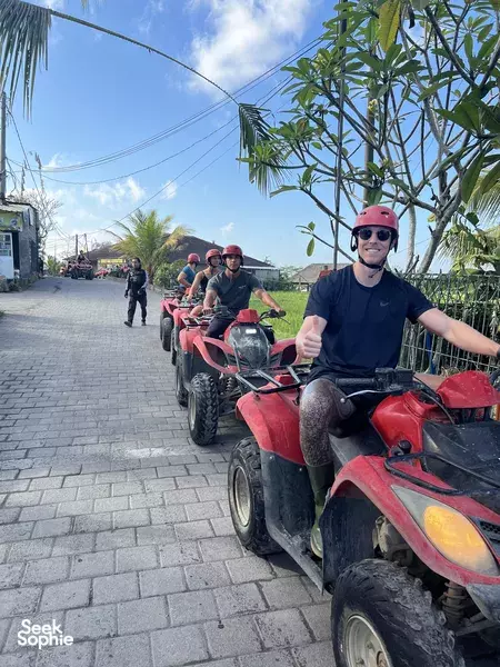 Helmed rider posing on a red ATV quad bike on a Bali paved street, with other machines and riders behind