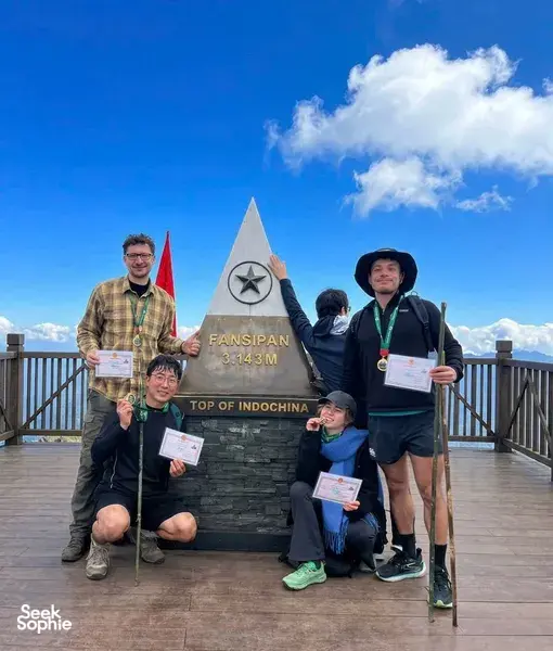 Trekkers posing at the Fansipan “Top of Indochina” sign on a 1-day eco trek in Sapa, with clouds above the summit