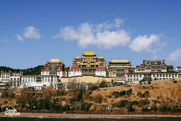 Traditional white-and-red temple complex with gilded rooftops above a riverbank in Shangri-La under a wide blue sky